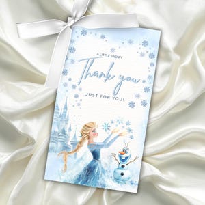 May include: A light blue gift tag with a white ribbon. The tag features a watercolor illustration of a princess and a snowman, with the words "A Little Snowy Thank You Just For You!" in a script font.