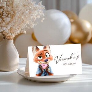 May include: A white place card with the text "Veronika's ICE CREAM" and a cartoon fox holding a pink heart-shaped donut. The card is on a white plate, with balloons in the background.