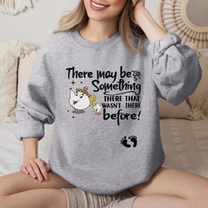 May include: A heather gray crewneck sweatshirt featuring a black graphic print of Mrs. Potts and Chip from Beauty and the Beast. The text reads, There may be Something THERE THAT WASN'T THERE before!, accompanied by a small black baby footprint icon.