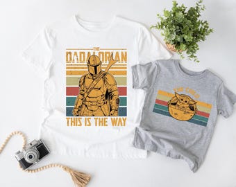 May include: A white t-shirt featuring a graphic of a armored character with the text THE DADALORIAN THIS IS THE WAY and a grey t-shirt featuring a graphic of a small creature in a pod with the text THE CHILD, both with retro striped backgrounds.