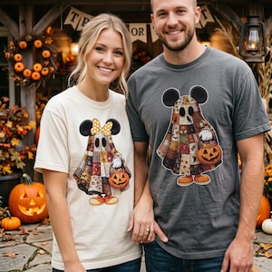 May include: A cream-colored t-shirt and a charcoal gray t-shirt, each featuring a graphic of a ghost wearing mouse ears and a patchwork-style sheet. The ghost holds a jack-o'-lantern candy bucket, and the cream shirt includes a yellow polka-dot bow.