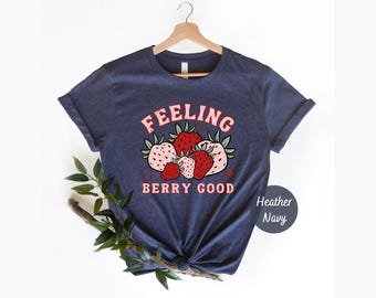 May include: A heather navy short-sleeve t-shirt featuring a graphic of five strawberries in shades of red and light pink. The text "FEELING BERRY GOOD" is printed in a light pink font above and below the strawberry illustration on the chest.