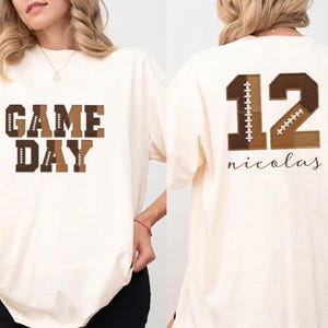 May include: An off-white short-sleeve t-shirt featuring the text GAME DAY in brown block letters on the front. The back displays the number 12 in brown with football lace patterns, accompanied by the name nicolas in a cursive brown script font.