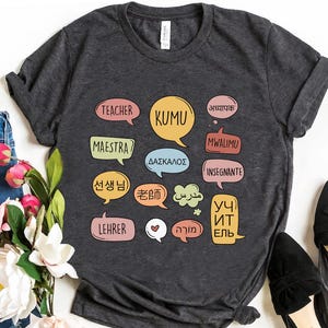 May include: A dark heather gray short-sleeve t-shirt featuring a graphic print of various colorful speech bubbles. Each bubble contains the word for teacher in a different language, including English, Spanish, German, Korean, Japanese, Greek, and others.