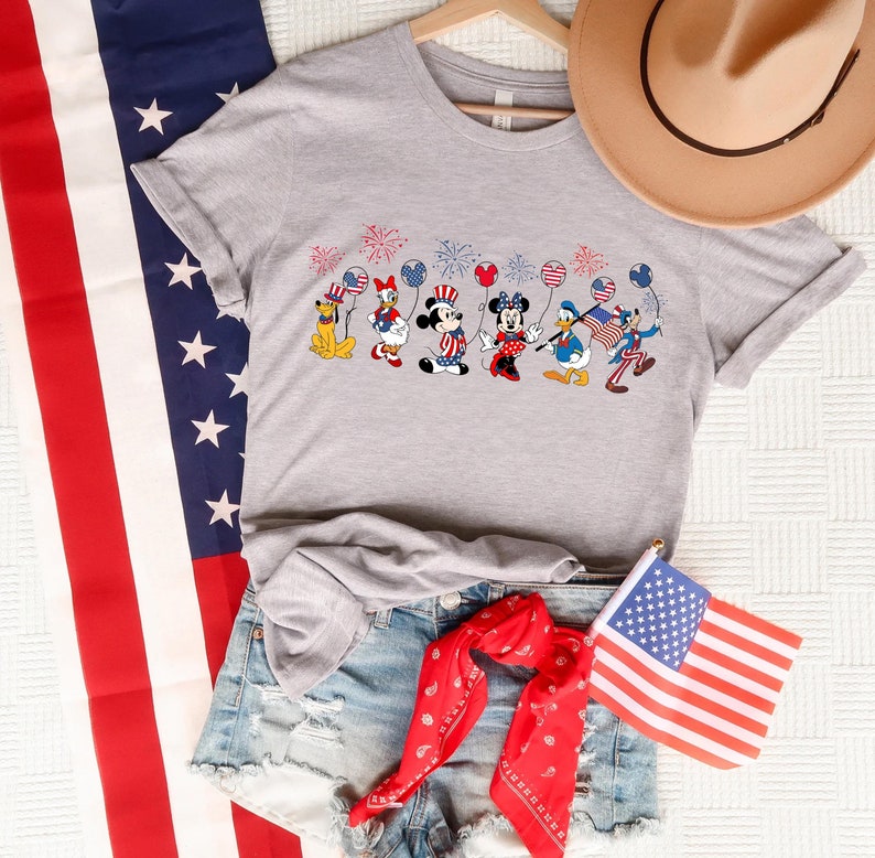 May include: Gray t-shirt featuring a patriotic design of Disney characters, including Mickey, Minnie, Goofy, Pluto, and Donald Duck, celebrating Independence Day with fireworks and balloons. The shirt is displayed with denim shorts, a red bandana, and an American flag, suggesting a summer or 4th of July theme.  The overall style is casual and festive.