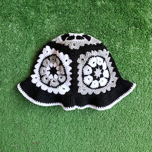 Pu&ograve; includere: Un cappello a secchiello in crochet nero e bianco con un motivo geometrico. Il cappello ha un'ampia tesa ed &egrave; realizzato con una combinazione di filato nero, bianco e grigio.