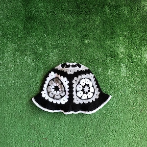Pu&ograve; includere: Un cappello a secchiello in crochet bianco e nero con un motivo geometrico. Il cappello &egrave; su uno sfondo di erba artificiale verde.
