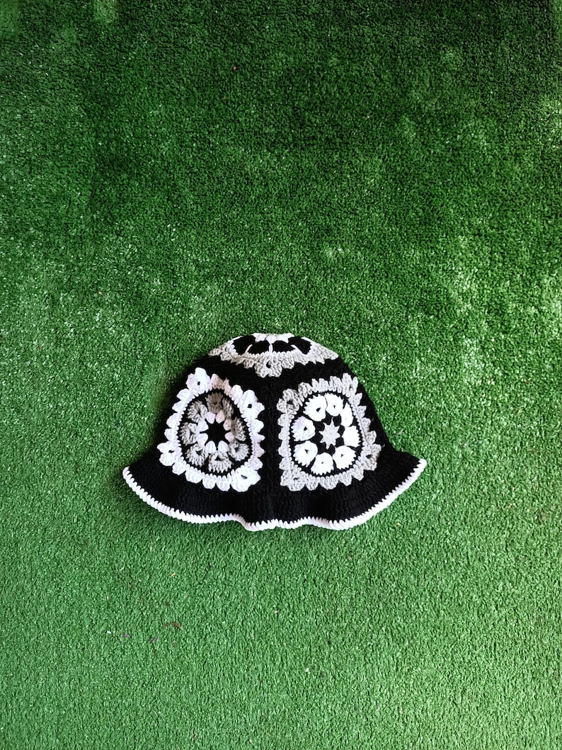 Pu&ograve; includere: Un cappello a secchiello in crochet bianco e nero con un motivo geometrico. Il cappello &egrave; su uno sfondo di erba artificiale verde.