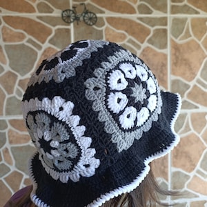 Pu&ograve; includere: Un cappello a secchiello in crochet nero e bianco con un bordo a scalare. Il cappello presenta un motivo ripetitivo di cerchi grigi e bianchi.