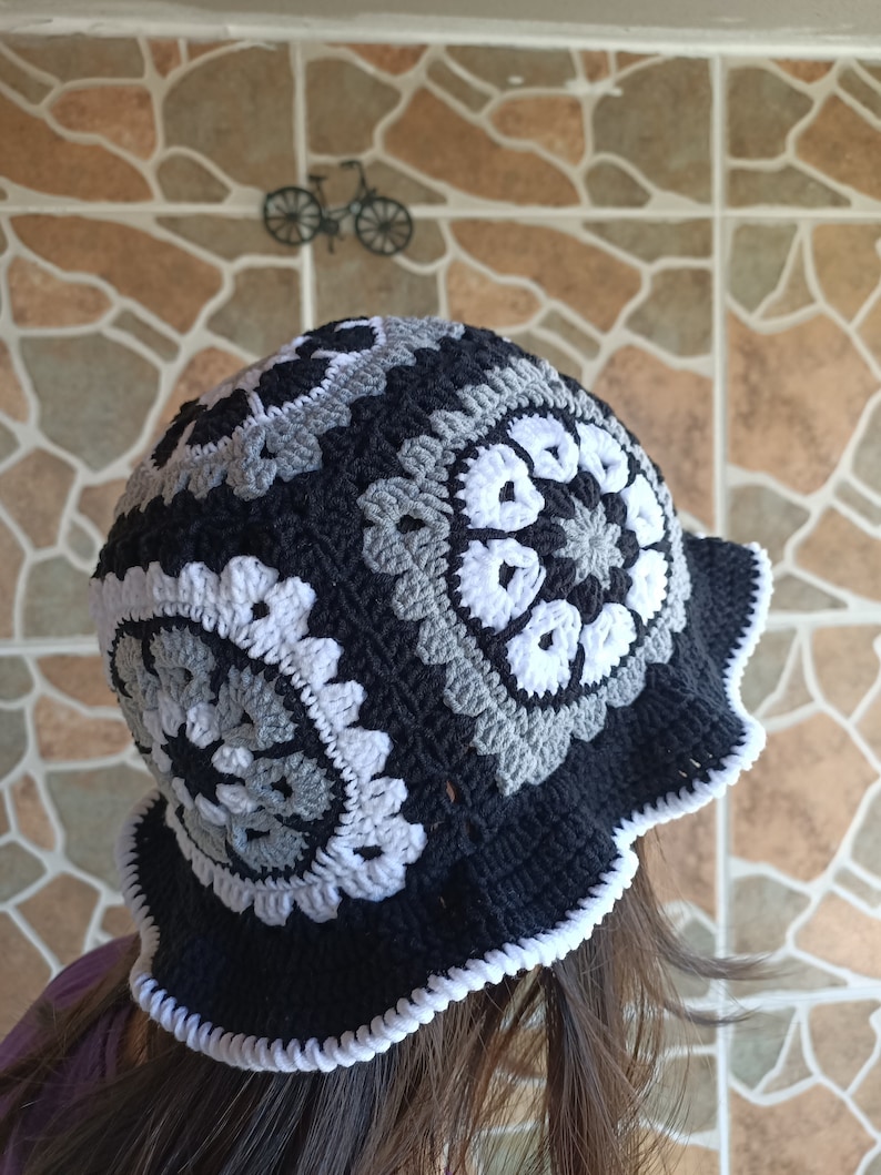 Pu&ograve; includere: Un cappello a secchiello in crochet nero e bianco con un bordo a scalare. Il cappello presenta un motivo ripetitivo di cerchi grigi e bianchi.