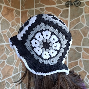Pu&ograve; includere: Un cappello a secchiello in crochet nero, bianco e grigio con un bordo in crochet bianco.