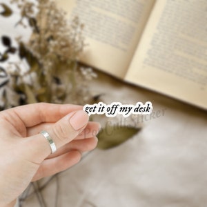 May include: A white sticker with the text "get it off my desk" in a black, handwritten-style font. The sticker is held by a person's hand, with a blurred background of a book and dried flowers.