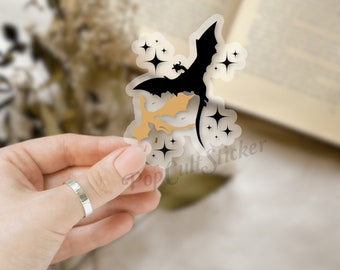 Könnte beinhalten: Ein transparenter Aufkleber mit einer schwarzen Silhouette eines Drachen, der mit einem kleineren Drachen in seinen Krallen fliegt. Der Aufkleber hat einen goldenen Umriss und Glitzer um die Drachen herum.