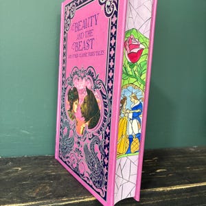Könnte beinhalten: Ein pinkfarbenes Hardcover-Buch mit dem Titel "Beauty and the Beast and Other Classic Fairy Tales". Das Cover zeigt Illustrationen der Charaktere. Der Buchrücken hat eine Buntglas-Illustration einer Rose und von Charakteren.