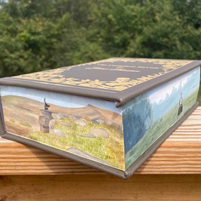 May include: A gray book with a gold design on the cover. The book is closed and the spine is visible. The book is sitting on a wooden surface. The book has a landscape scene on the front cover with a mountain in the background and a stone structure in the foreground.