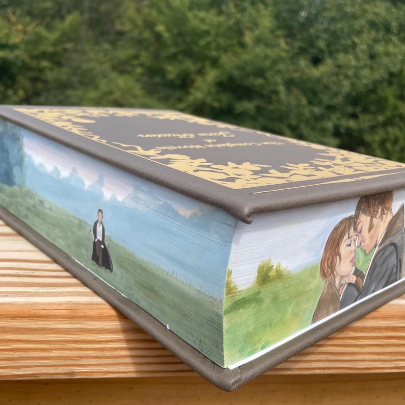 May include: A gray book with gold lettering on the spine that reads "Pride and Prejudice" by Jane Austen. The book is painted with a scene from the novel on the front and back covers. The front cover shows a couple kissing in a field, while the back cover shows a man walking in a field.