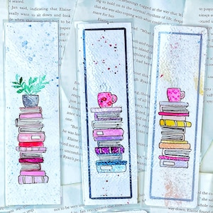 Puede incluir: Tres marcadores de acuarela con pilas de libros con una taza de café en la parte superior. Los marcadores están sobre un fondo blanco con una acuarela de color azul claro y rosa.