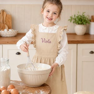 May include: A beige apron with ruffled shoulder straps and the name Viola embroidered in purple thread on the chest. The apron is worn over a white long-sleeved shirt with a collar, featuring a whisk and a large mixing bowl in the foreground.