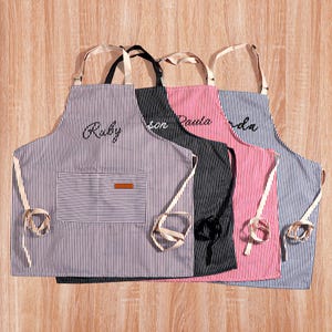 May include: Four striped aprons in blue, black, and red. Each apron features a pocket and the names "Ruby", "son", "Paula", and "da" embroidered on them. The aprons have cream-coloured ties and neck straps.
