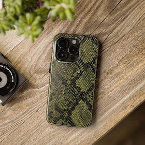 May include: A green and black snakeskin-patterned phone case. The case is on a wooden surface. The phone case has a camera with four lenses. A black camera and green plant are in the background.