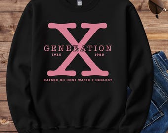 May include: Black sweatshirt featuring a pink "X" graphic with the word "GENERATION" above the years "1965" and "1980". The text "RAISED ON HOSE WATER & NEGLECT" is below. Sunglasses and denim jeans are also visible in the image.