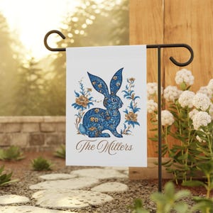 May include: A white garden flag with a blue and gold rabbit design. The rabbit is surrounded by flowers and the text "The Millers" is printed below the rabbit.
