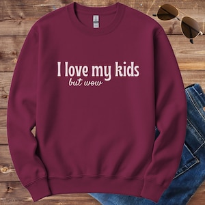 May include: A maroon sweatshirt with the text "I love my kids but wow" in white. The sweatshirt is displayed on a wooden surface with sunglasses and a pair of jeans.