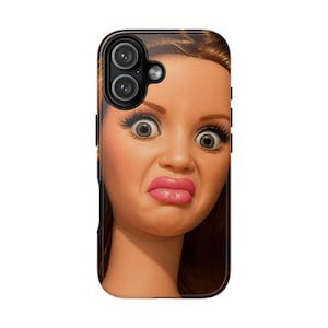 May include: Black phone case featuring a close-up of a doll's face with a surprised expression. The doll has large eyes, long eyelashes, and pink lips. The case has a black border and a dual-lens camera cutout.