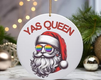 May include: A white ceramic Christmas ornament with a red and white Santa Claus wearing rainbow sunglasses and a beard. The ornament has the text "YAS QUEEN" in red letters.