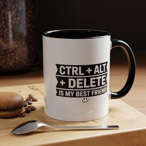 May include: White ceramic mug with a black interior and handle. The mug features the text "CTRL + ALT + DELETE IS MY BEST FRIEND" in black, with a smiley face. The mug is on a wooden surface.