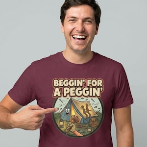 Könnte beinhalten: Burgunderfarbenes T-Shirt mit dem Text "Beggin' For A Peggin'" über einer Camping-Grafik. Die Grafik zeigt ein Zelt, einen Hammer und andere Campingausrüstung. Die Person im Bild zeigt auf die Grafik.