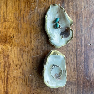 Puede incluir: Dos conchas de ostra utilizadas como joyeros sobre una superficie de madera. Una contiene pendientes turquesa y un fragmento de concha. La otra contiene un anillo de plata con una piedra oscura y piedras laterales más pequeñas. Las conchas tienen bordes pintados de dorado.