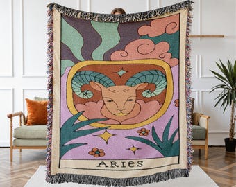Könnte beinhalten: Eine gewebte Wandteppichdecke mit einem Widder-Tierkreiszeichen-Design. Die Decke zeigt eine Widder-Illustration in einem rechteckigen Rahmen, umgeben von Pastellfarben und floralen Akzenten. Das Wort "ARIES" ist unten aufgedruckt. Die Decke hat einen Fransenrand.