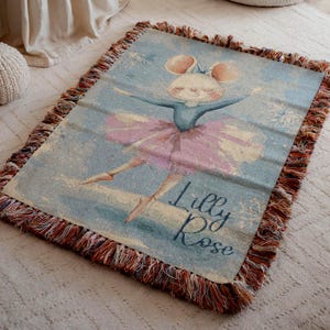 Puede incluir: Una manta tejida rectangular con fondo azul claro y una ilustración central de un ratón con tutú rosa y zapatillas de ballet. El nombre Lilly Rose está impreso en letra cursiva azul oscuro. Los bordes tienen flecos de varios colores.