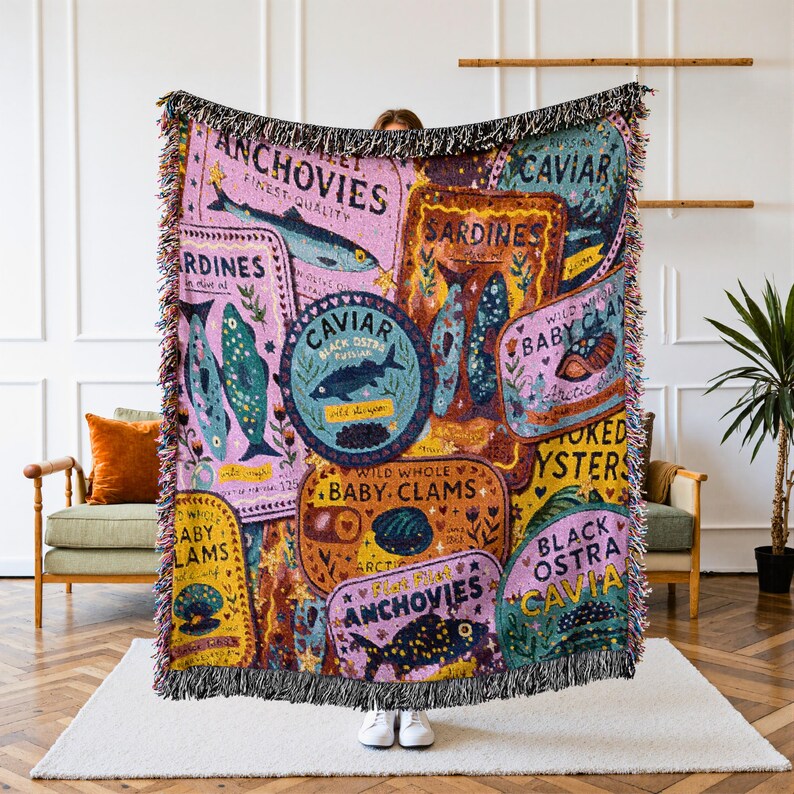Vintage-style woven tapestry blanket featuring colorful sardine, anchovy, caviar, and clam labels,  Blanket is being held up by a person in a living room setting.