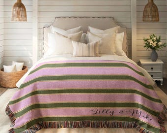 May include: A striped blanket in shades of lavender and olive green, draped over a bed. The blanket features horizontal stripes and a fringed edge. The names "Gilly & Mike" are embroidered on the blanket.
