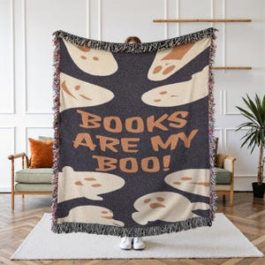 Peut inclure: Une couverture tissée gris foncé avec un motif répété de fantômes beiges. L'expression "BOOKS ARE MY BOO!" est imprimée en lettres blocs beiges. La couverture a une bordure frangée.