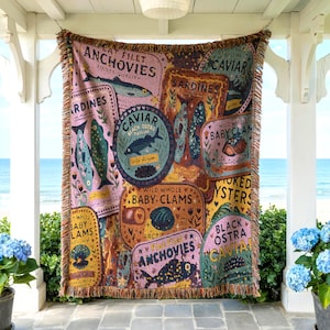 Vintage-style woven tapestry blanket featuring colorful sardine, anchovy, caviar, and clam labels, displayed hanging on a seaside porch with ocean views and coastal decor.