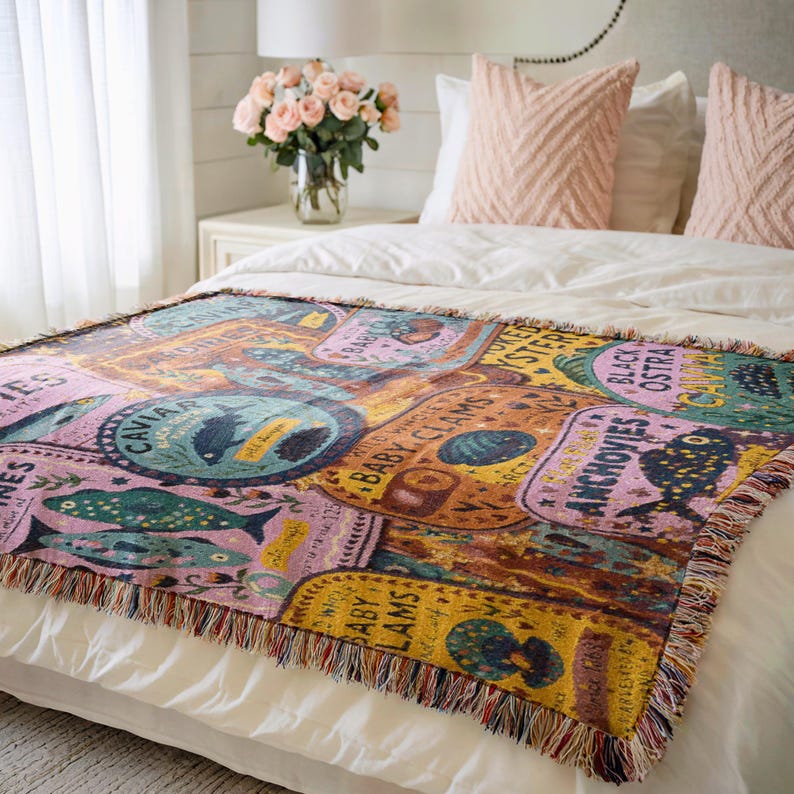 Vintage-style woven tapestry blanket featuring colorful sardine, anchovy, caviar, and clam labels in a collage design.  Blanket is being shown as bedding