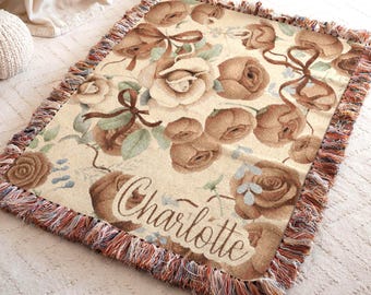 May include: A rectangular woven throw blanket featuring a cream background with a pattern of brown roses, green leaves, and brown ribbons. The name Charlotte is written in a cursive script across the lower section. The edges have a fringed border.