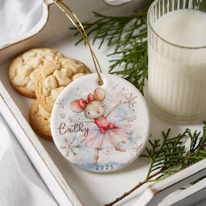 Könnte beinhalten: Runder Weihnachtsbaumschmuck aus Keramik mit einer tanzenden Mäuseballerina in einem rosa Tutu und einer roten Schleife. Der Schmuck trägt den Namen "Cathy" und das Jahr "2025". Er wird von einem goldenen Faden gehalten. Kekse und ein Glas Milch sind im Hintergrund.