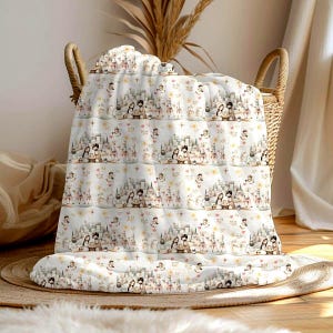 May include: A white baby blanket featuring a repeating pattern of a fairytale scene, including castles, angels, and couples. The blanket is folded in a wicker basket, with a neutral background.