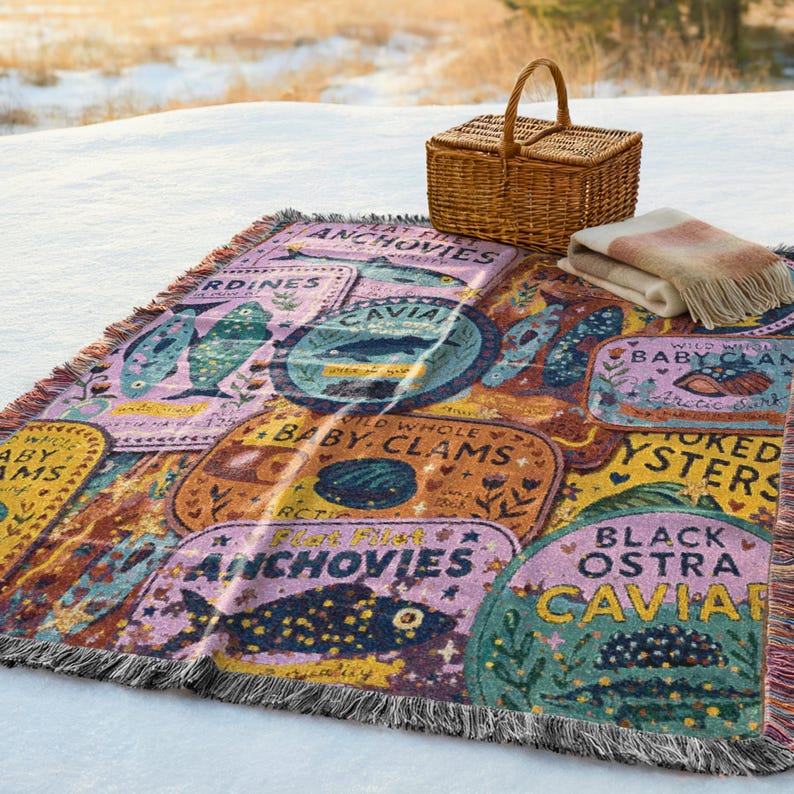 Vintage-style woven tapestry blanket featuring colorful sardine, anchovy, caviar, and clam labels in a collage design.  Blanket is being shown as a picnic blanket in a beach setting.