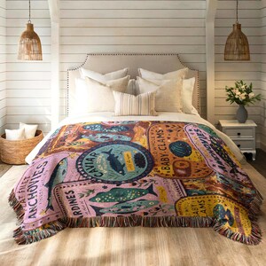 Vintage-style woven tapestry blanket featuring colorful sardine, anchovy, caviar, and clam labels in a collage design.  Blanket is being shown as bedding in a foot of a bed.