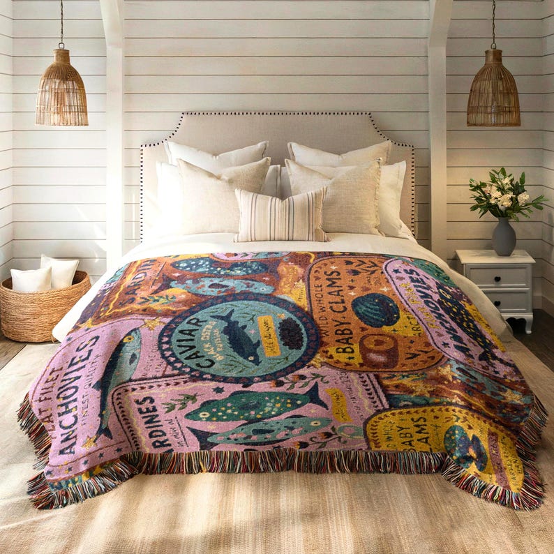 Vintage-style woven tapestry blanket featuring colorful sardine, anchovy, caviar, and clam labels in a collage design.  Blanket is being shown as bedding in a foot of a bed.
