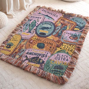 Vintage-style woven tapestry blanket featuring colorful sardine, anchovy, caviar, and clam labels in a collage design.  Blanket is shown resting on a carpeted floor