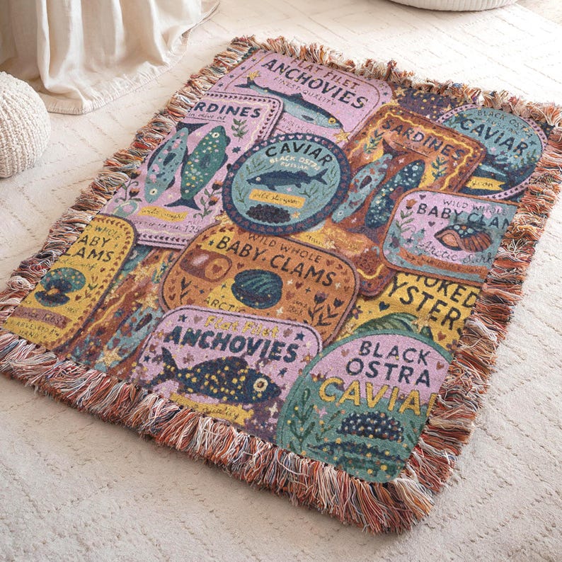 Vintage-style woven tapestry blanket featuring colorful sardine, anchovy, caviar, and clam labels in a collage design.  Blanket is shown resting on a carpeted floor