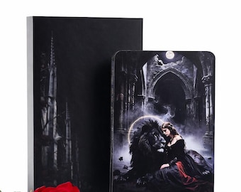 May include: A deck of tarot cards featuring the Strength card with a black lion and a figure in a black dress, housed in a black box with a gothic cathedral illustration, accompanied by a single vibrant red rose with green leaves.
