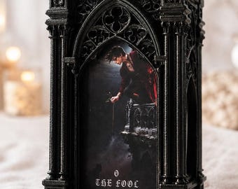 May include: A black, rectangular, gothic-style frame featuring intricate architectural details like columns and arches. Inside the frame is an illustration of a person on a balcony holding a flower, with the text 0 THE FOOL printed at the bottom.