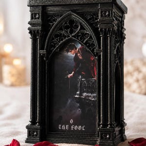 Peut inclure: Un cadre noir rectangulaire de style gothique avec des détails architecturaux complexes tels que des colonnes et des arches. À l'intérieur se trouve une illustration d'une personne sur un balcon tenant une fleur, avec le texte 0 THE FOOL.